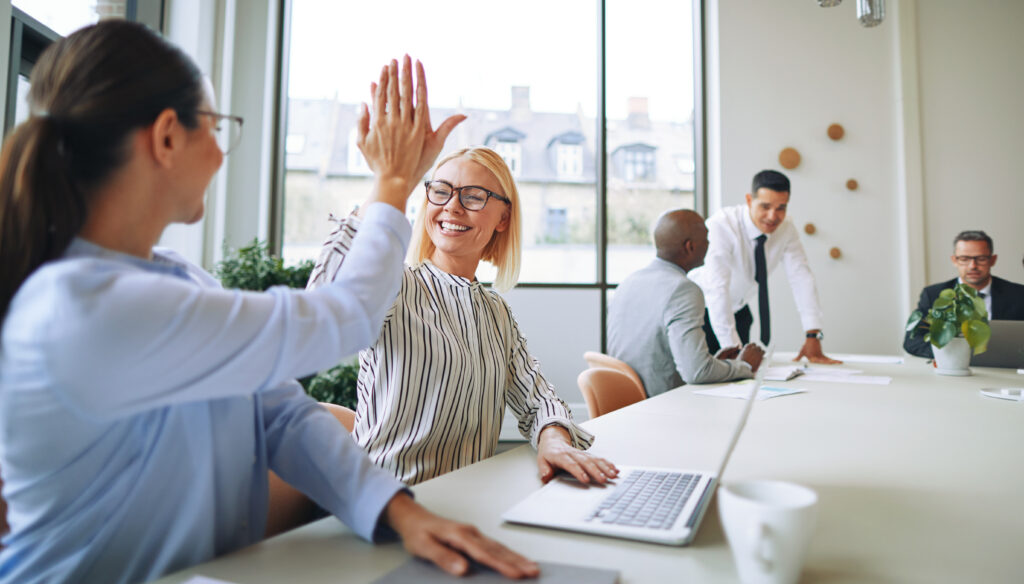 Two young businesswomen smiling and high fiving together while sitting at a boardroom table with colleagues in the background