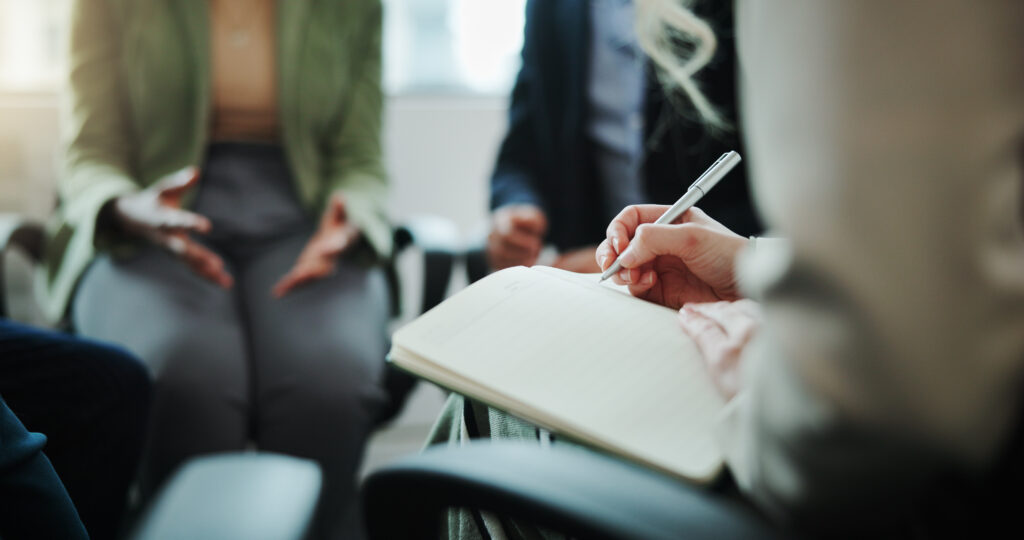 Business people, hands and writing with book in meeting for support group, therapy session or counseling. Closeup, hr or taking notes with team for company dispute or conflict resolution at workplace.