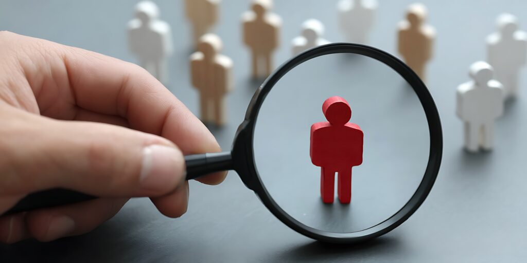 A hand uses a magnifying glass to focus on a single red figure among many others, symbolizing talent search and recruitment.