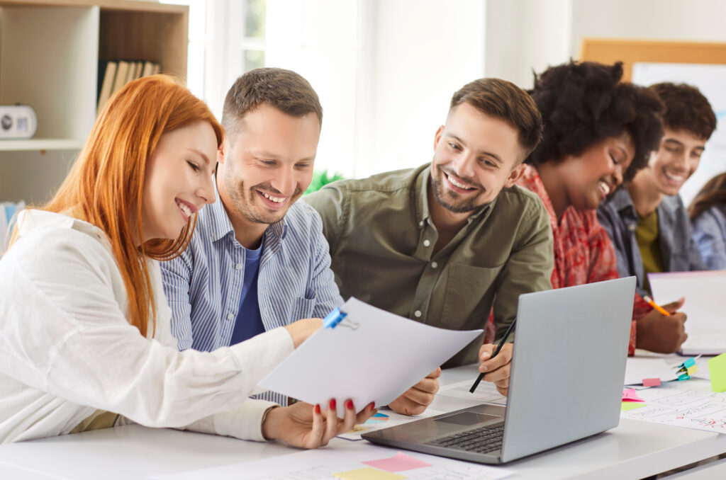 Team of happy business people working together on paperwork and a laptop during a meeting in the office. The group engages in conversation and collaboration, discussing business matters.