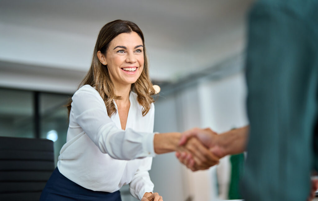 Happy mature professional business woman handshaking partner in office. Smiling female hr manager of middle age making successful deal with partner shaking hand of client at work standing at meeting.